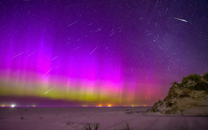 Purple and green aurora over a dark landscape with meteors overhead.